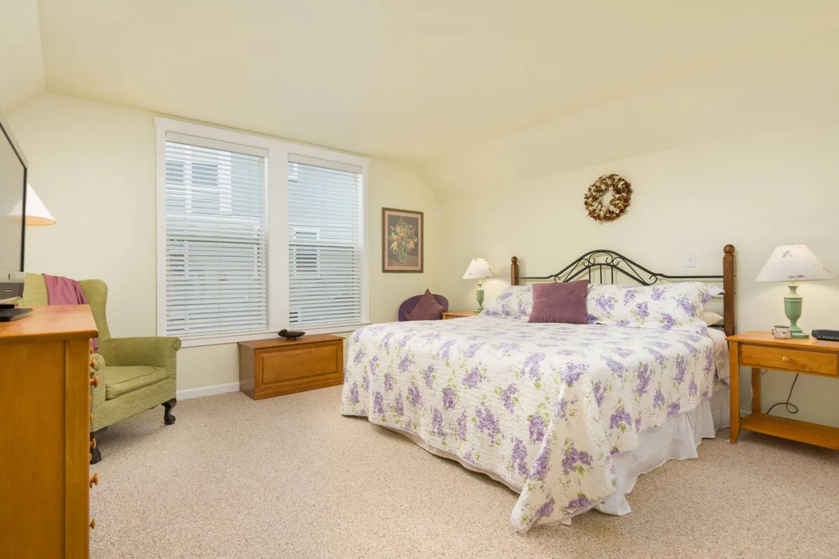A serene bedroom scene featuring a bed with floral bedding, a cozy green chair for reading, and wooden nightstands. Natural light streams in, creating a calm and restful atmosphere perfect for unwinding after a long day.