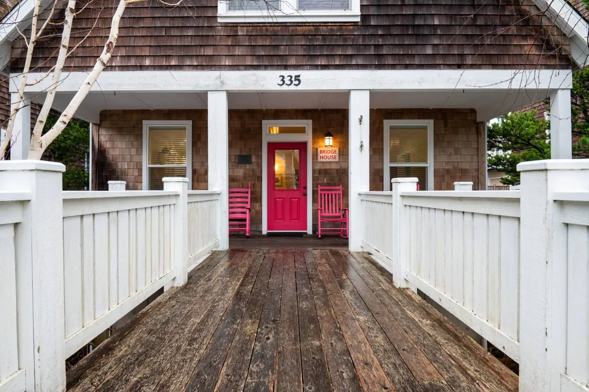 The image showcases the exterior of The Bridge House in Depoe Bay, Oregon, featuring a charming wooden walkway leading to a bright pink front door. Red rocking chairs sit invitingly on the porch, suggesting a cozy spot to relax and take in sunset views. The scene radiates warmth and hospitality, set against the coastal backdrop.