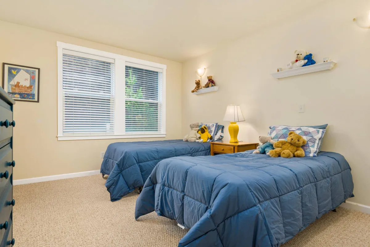 A welcoming twin-bedded room adorned with stuffed animals and playful touches, including a nightstand with a lamp and framed artwork. The scene evokes childhood joy and comfort, ideal for families with kids or groups of friends.