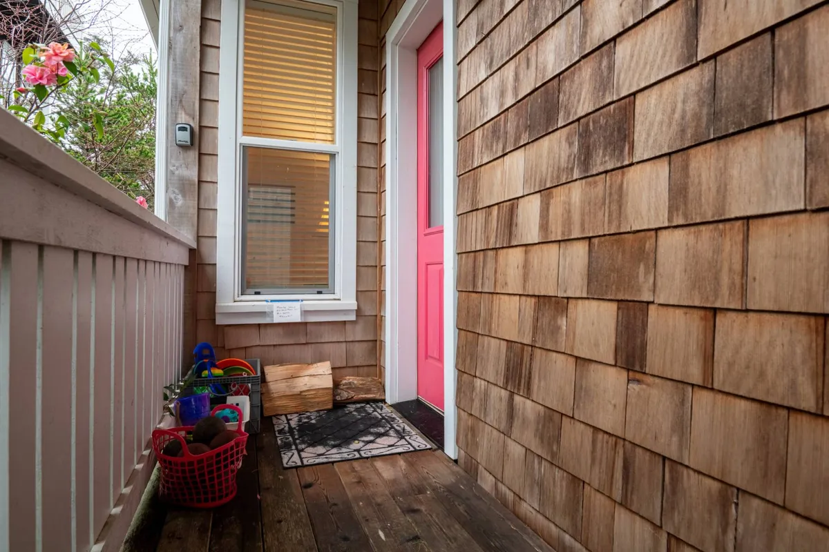An exterior scene featuring a home with a cheerful pink door, wooden shingles, a decorative mat, and baskets of colorful toys. The porch area is welcoming, suggesting both relaxation and playful outdoor activities.