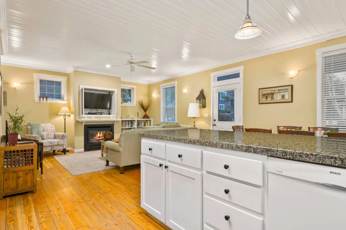 This interior image beautifully captures the open-plan living and kitchen area, showcasing a cozy seating arrangement near a gas fireplace, a spacious kitchen island, and large windows that allow abundant natural light. The visible connection between the seating, kitchen, and dining areas highlights the communal design and inviting atmosphere.