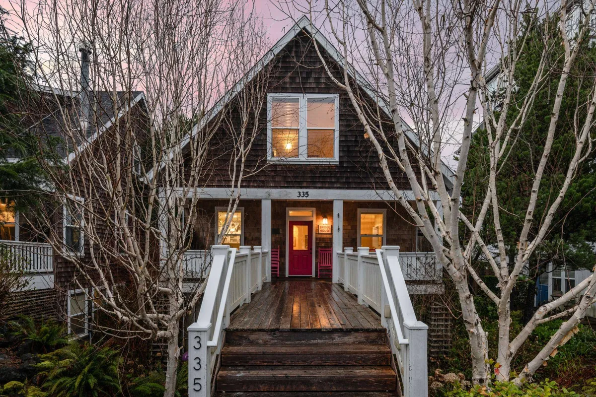 A vibrant pink front door anchors a welcoming exterior, framed by large windows that glow with warm interior light. Leafless trees surround the entryway, and the shady porch invites guests to gather, sip coffee, or enjoy sunset conversations.