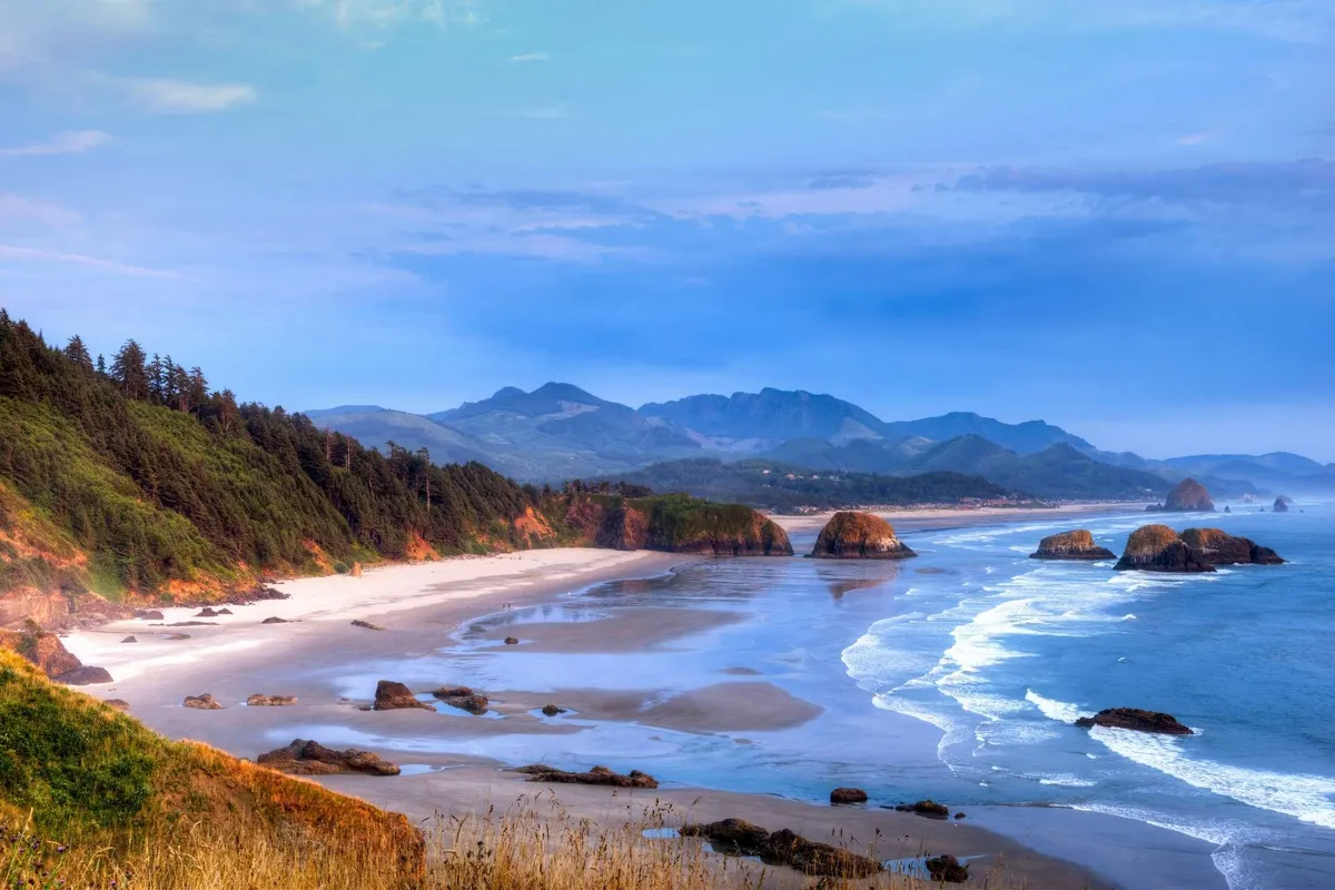 The sweeping view of an expansive sandy beach, punctuated by dramatic rock formations and the backdrop of distant mountains. Ocean waves roll gently onto shore, inviting exploration and reflection.