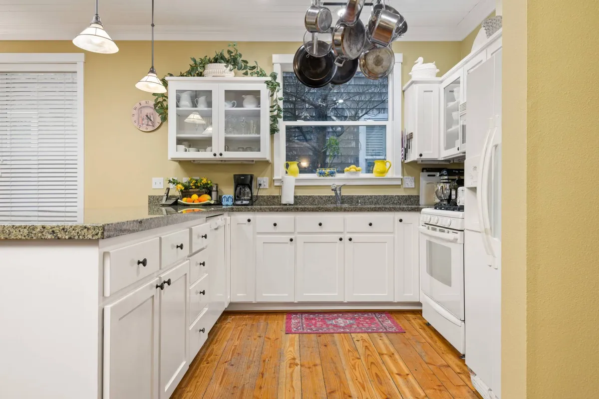 A well-lit gourmet kitchen scene, with a granite countertop, white cabinetry, a European-style hanging rack with pots, colorful dishware, and a window flooding the space with natural light. The kitchen is both functional and inviting, encouraging culinary creativity and social interaction.