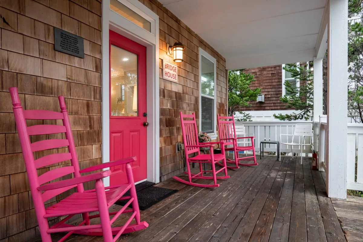 A close-up exterior shot highlighting the porch’s pink rocking chairs, the bright front door, and classic wooden siding. The scene exudes cheerfulness and comfort, suggesting an ideal spot for relaxing or socializing with friends. The setting’s brightness and arrangement of furniture make the porch feel like a true outdoor living room.
