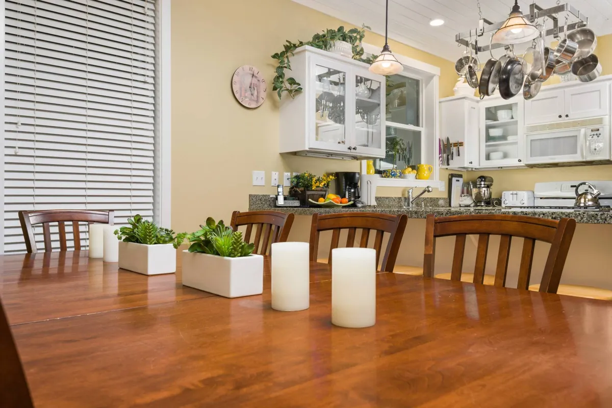 A warm, welcoming dining area featuring a large wooden table set with candles and surrounded by comfortable seating. Hanging pots and pans above hint at the proximity of the kitchen, while succulent plants add a touch of freshness. The table is clearly built for group gatherings and shared meals.