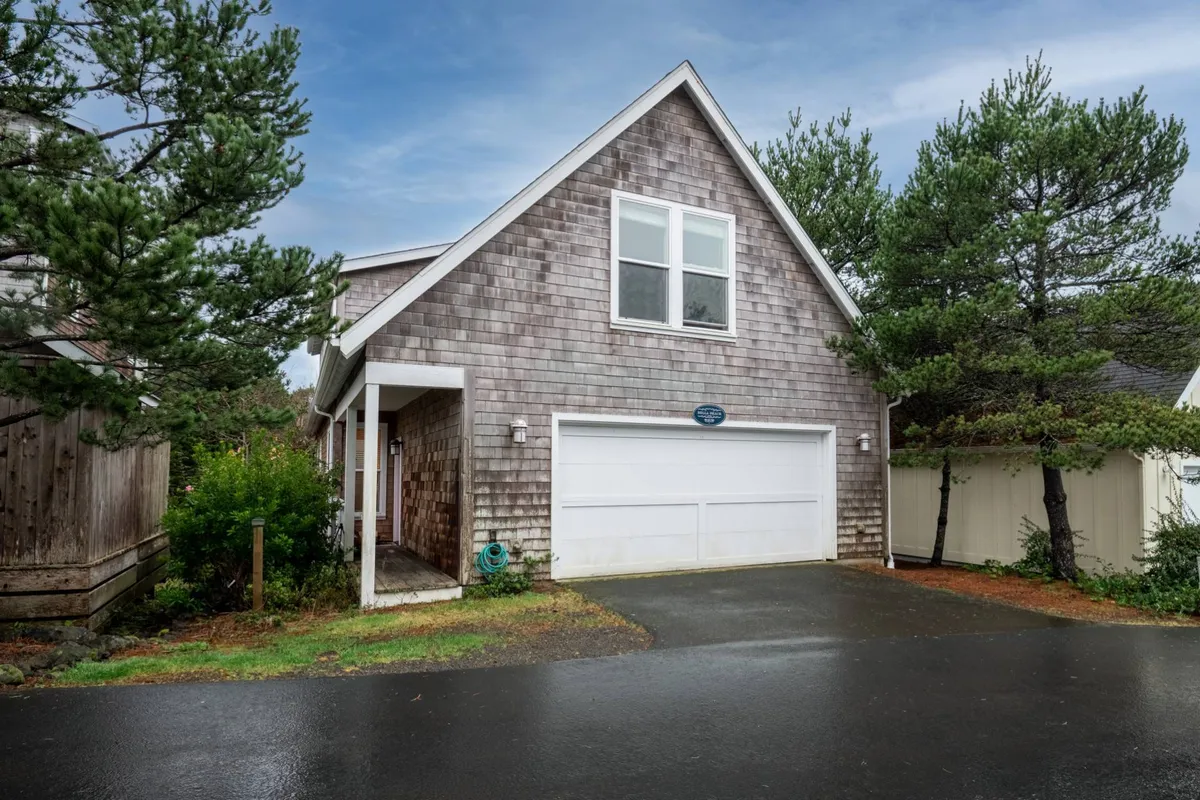 A wider exterior view showing the home’s shingle siding, double windows, garage door, and a portion of the driveway. Though less focused on the porch itself, the image hints at the porch area and highlights the home’s peaceful, tree-lined setting, reinforcing the sense of privacy and connection with nature. This broader perspective situates the porch in its tranquil Bella Beach environment.