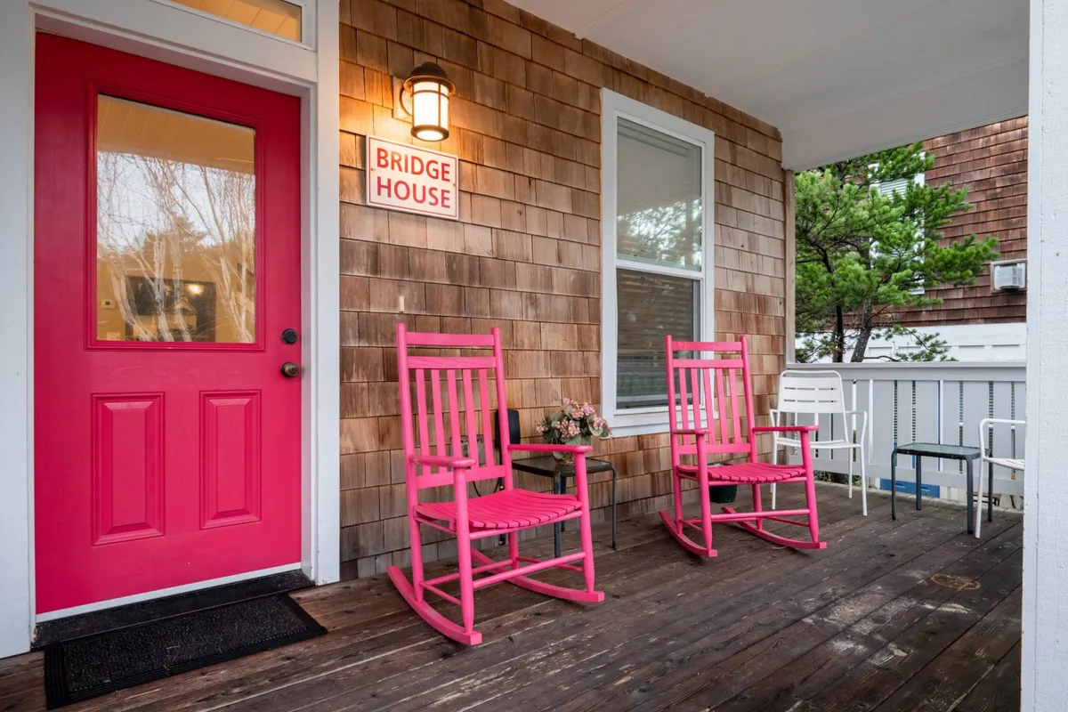 A spacious front porch featuring cheerful pink rocking chairs, a bright pink door, the 'BRIDGE HOUSE' sign, wooden siding, and a welcoming planter with vibrant flowers. The setting is shaded by surrounding trees, providing a sense of privacy and tranquility. The arrangement and decor invite relaxation and socialization.