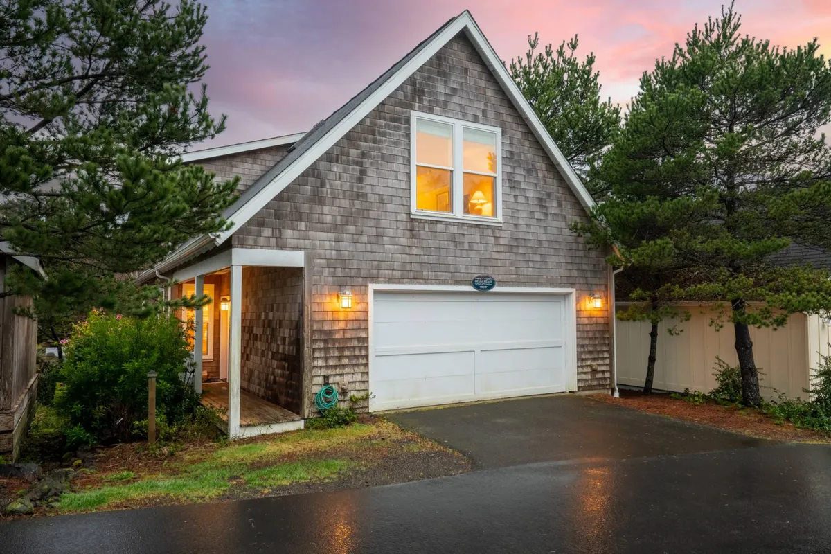 The home’s textured wooden exterior glows with the light from inside, while the driveway and garage area add context to the property’s setting. The porch and illuminated windows hint at cozy gatherings and the warmth awaiting guests after a day of exploration.