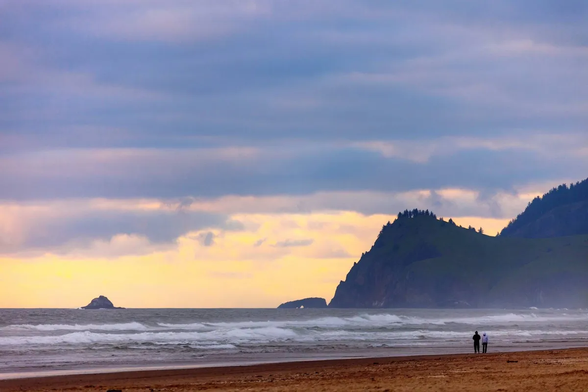 A tranquil scene featuring gentle ocean waves, distant cliffs, and a couple walking hand in hand along the sandy beach. The soft lighting and open space create a sense of relaxation, peace, and the playful exploration of the shore.