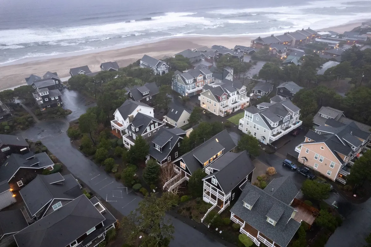 A sweeping view captures a stretch of sandy beach bordered by ocean waves, with charming houses in the background and a tree line framing the shoreline. The image conveys the expanse of the beach, the neighborhood’s proximity to the coast, and the inviting natural environment, ideal for walks, relaxation, and ocean fun.